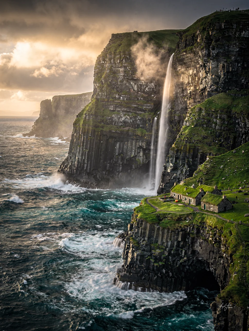 Dramatic green turf-roofed stone cottages perched beneath monolithic sea cliffs in the Faroe Islands